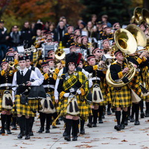 The College of ۿ۴ý marching band marches in uniform with alumni and families trailing behind.